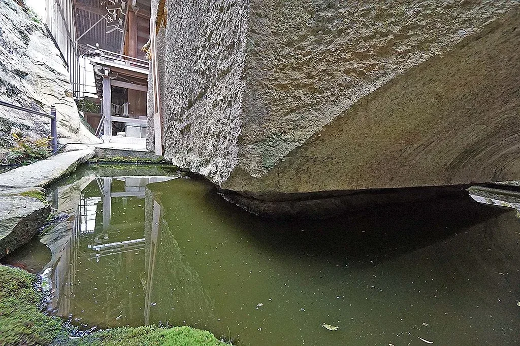 Pogled iz blizine na masivni kameni monolit sa precizno obrađenim ivicama, koji se nadvija nad bazenom mirne vode. Odraz kamena i neba stvara impresivnu refleksiju, naglašavajući osećaj spokoja