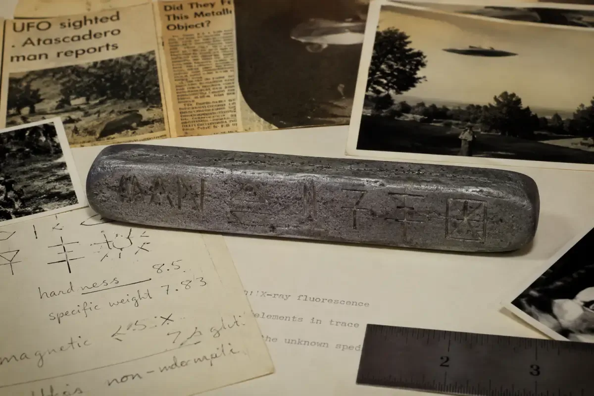 AI ilustracija metalne poluge s ugraviranim natpisom "MAN" i nepoznatim simbolima na istraživačkom stolu okruženom dokumentima i novinskim isječcima iz navodnog UFO slučaja iz 1970-ih.