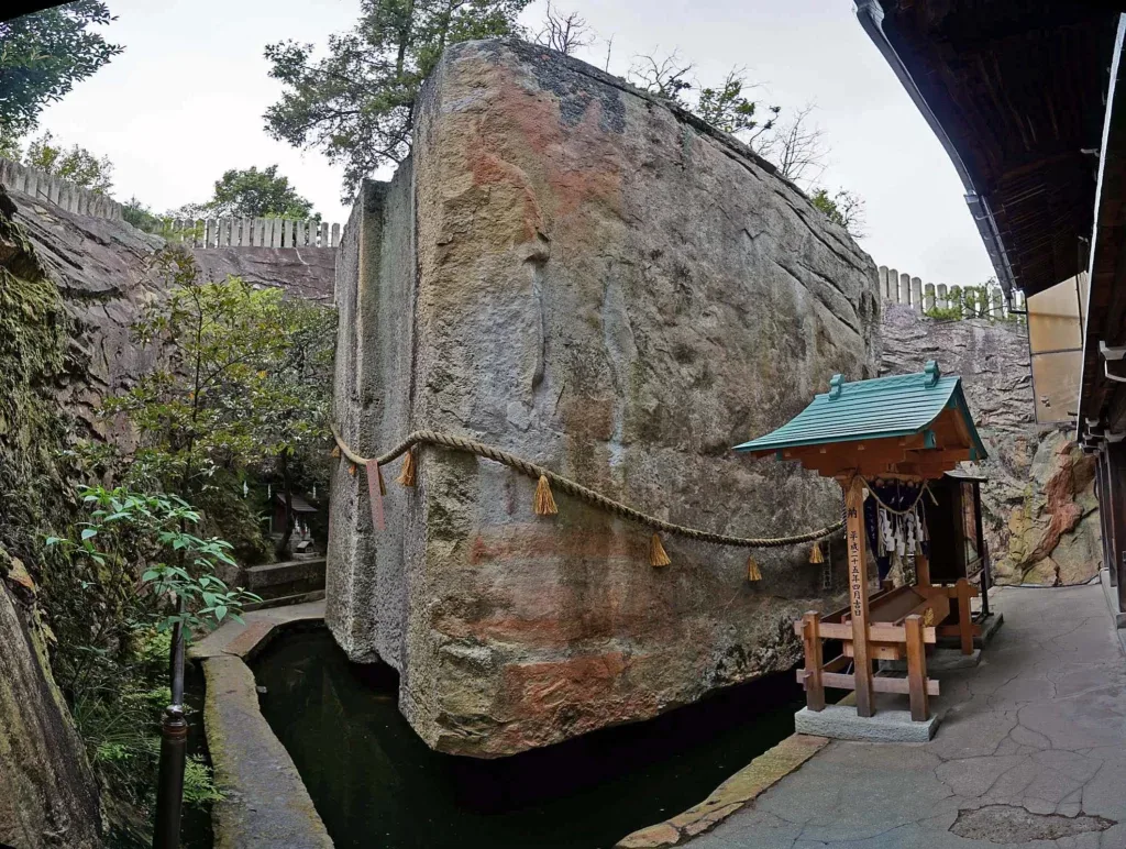 Detaljan pogled na kolosalni kameni monolit obmotan svetim konopcem sa resama, postavljen u podnožju kamenoloma. Pored stene nalazi se tradicionalno japansko svetilište sa tamnoplavom zavesom i malim drvenim detaljima